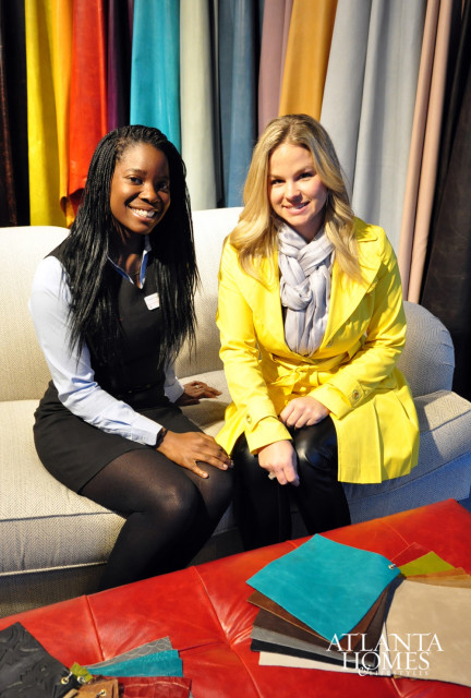 Two women sitting on a sofa in front of a display of colorful leather swatches. The woman on the left has dark braided hair and wears a black vest over a light blue shirt. The woman on the right has blonde hair and wears a yellow coat and grey scarf.