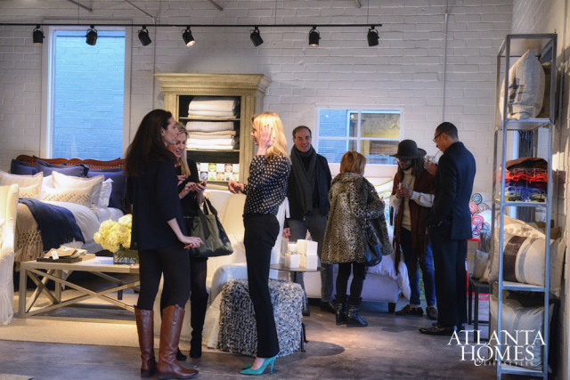People gather in a showroom featuring a bed with navy blue bedding, a wooden cabinet filled with folded linens, and a metal shelving unit displaying pillows and blankets.