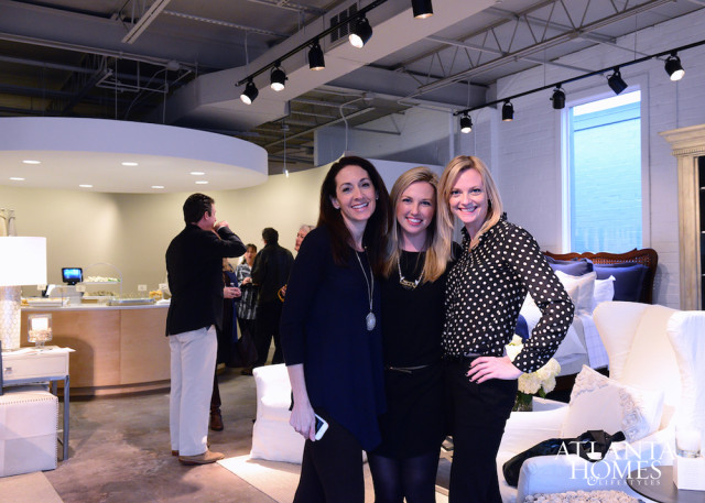 Three women smiling at a reception with a catering station and furniture displays in the background.