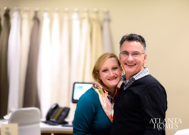 A smiling couple poses for a photo in front of a wall of fabric samples. The woman wears a teal top and a colorful scarf. The man wears a black sweater over a plaid shirt.