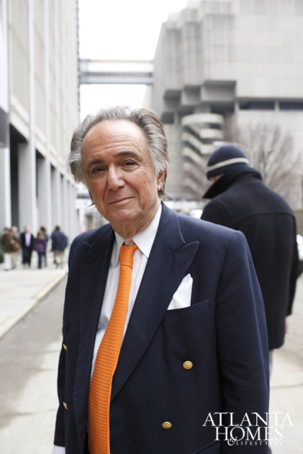 Man in a navy blue suit with an orange tie and white pocket square, standing outside with buildings in the background.