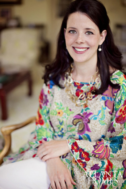 Woman with dark hair and green eyes smiling, wearing a colorful floral print top and a chunky gold necklace.