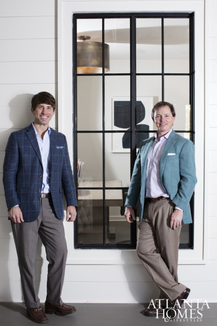 Two men in suits stand in front of a large window with a grid pattern. The man on the left wears a blue plaid jacket and the man on the right wears a teal jacket.
