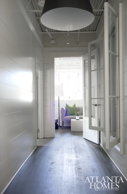 Hallway with white shiplap walls, dark wood floors, and a large black pendant light. A doorway opens to a sitting area with a lavender sofa, green pillow, and white coffee table.