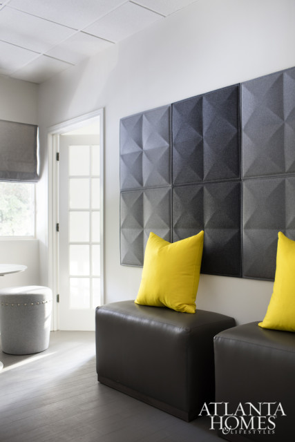Modern waiting area with dark leather ottomans topped with bright yellow pillows, a geometric dark gray wall paneling, and a grey upholstered ottoman near a window.