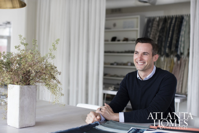 Man in navy sweater and collared shirt smiling, seated at a table with a vase of dried flowers and fabric swatches. Atlanta Homes & Lifestyles logo in the lower right corner.