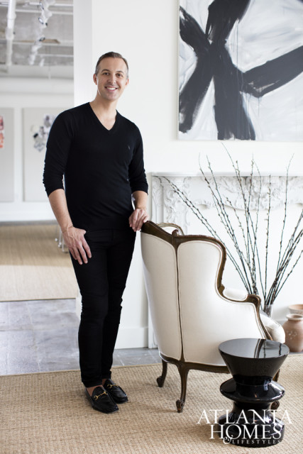 Man in black v-neck shirt and black pants standing next to a white armchair with ornate wooden trim.