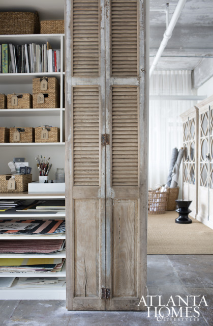 White shelving unit with woven baskets and books next to weathered wooden louvered doors. In the background, a black stool sits on a rug near cabinets.