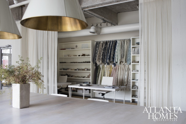 Interior design studio with fabric swatches hanging on wall, white shelves with books and samples, and a large table in the foreground.