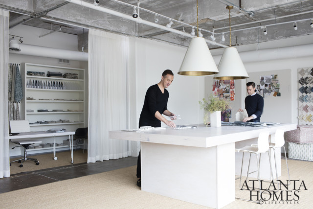 Interior design studio with two men working at a large white table, shelves of fabric samples, and a large white pendant light overhead.