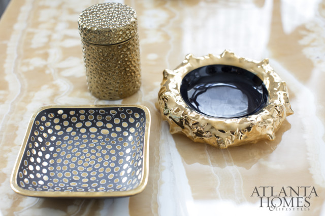 Gold textured jar, black and gold perforated square dish, and a gold organic-shaped bowl with a dark interior sit on a marble surface.