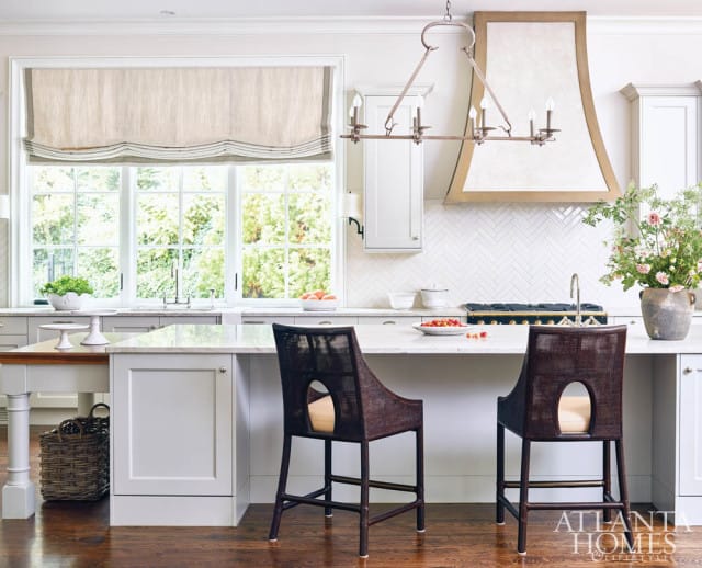 White kitchen with marble island, herringbone tile backsplash, range hood with decorative trim, and two dark woven bar stools.
