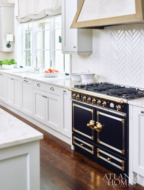 White kitchen with white herringbone tile backsplash, a dark blue range with brass accents, and a brass range hood. White cabinetry and a marble countertop are visible.