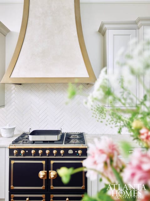 Black range with brass accents and a white herringbone tile backsplash topped by a textured stone range hood with brass trim.