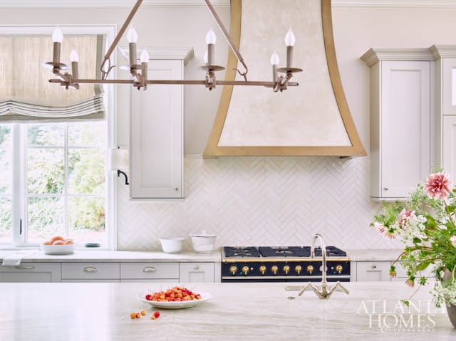 White kitchen with herringbone tile backsplash, brass range hood, and a dark blue range. A bowl of cherries sits on the counter in the foreground.