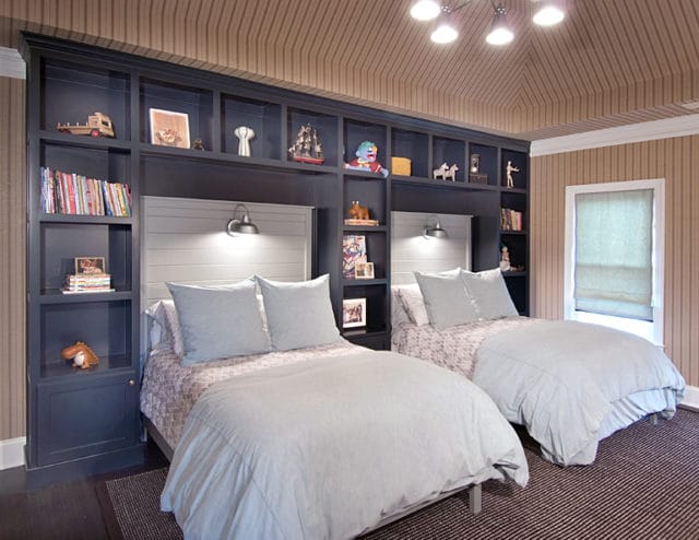 Children's bedroom with two beds, navy built-in bookshelves, light blue bedding, and striped wallpaper.