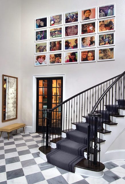 Entryway with a checkered marble floor, a dark wood staircase with a gray runner and black spindles, a mirrored wall hanging, and a grid of framed family photos on the wall.