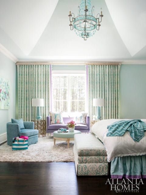 Bedroom with light blue walls, a vaulted ceiling with a blue chandelier, patterned curtains, a purple sofa, and a bed with a teal chevron blanket.