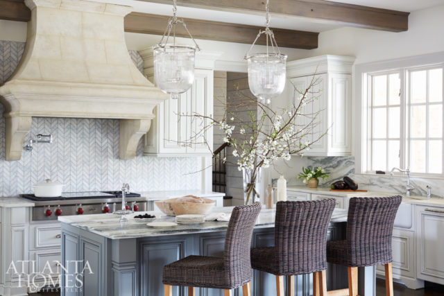 The kitchenjuxtaposes varying textures rather than contrasting colors. The Carrara marble backsplash—laid in a herringbone pattern next to the stone hood, wooden beamed ceiling, and veiny Vermont marble countertops—brings a soft, subtle contrast to the space.