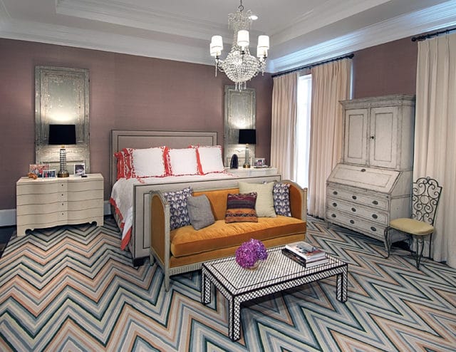 Master bedroom with a king-size bed, orange velvet sofa, chevron rug, and antique secretary desk. Walls are a muted lilac with distressed mirror panels.