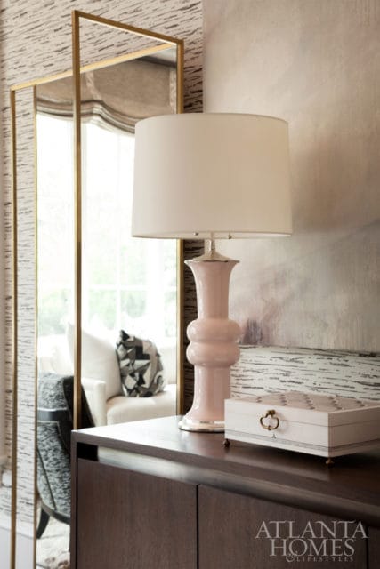 Pink ceramic lamp with white shade on a dark wood dresser, next to a decorative box and a tall gold-framed mirror reflecting a sofa.