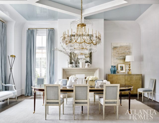 Elegant dining room with a glossy pale blue coffered ceiling, a large crystal chandelier, a dark wood dining table with upholstered chairs, a fireplace, and a gold credenza.