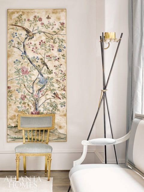 Gilded chair with light blue upholstery in front of a large tapestry depicting birds and flowers. A modern floor lamp stands to the right.