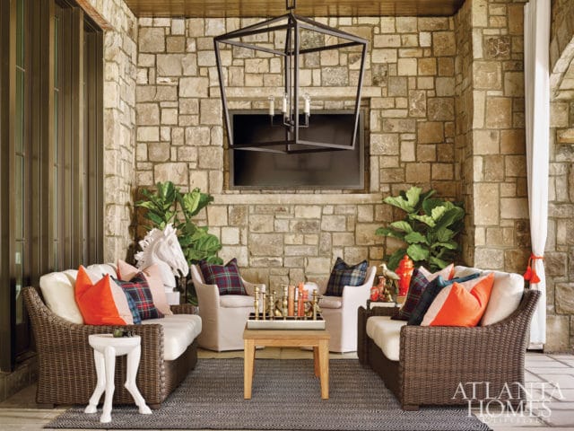 Covered patio with stone walls, wicker sofas, plaid and orange pillows, and a modern black lantern chandelier.