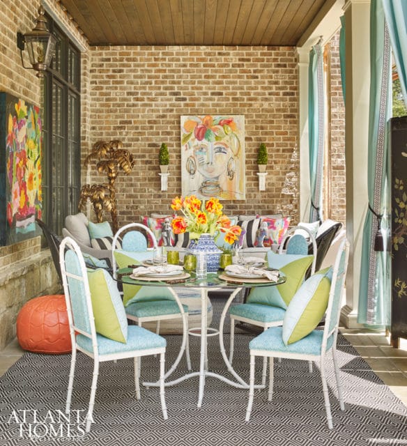 Covered patio with brick walls, wood ceiling, and a glass-top dining table set for four. Features include colorful pillows, artwork, and teal curtains.