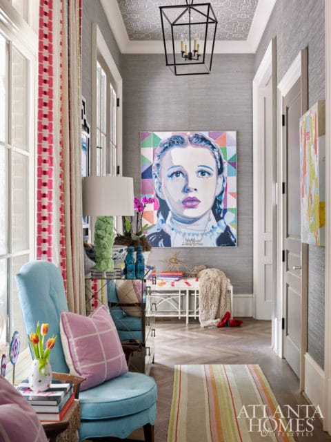 Hallway with gray grasscloth wallpaper, a large portrait of Judy Garland, a blue armchair with pink checkered pillow, and a geometric patterned ceiling