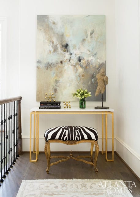 Hallway console table with marble top and gold frame, topped with abstract art, books, and a green vase of flowers. A zebra-print bench sits in front.