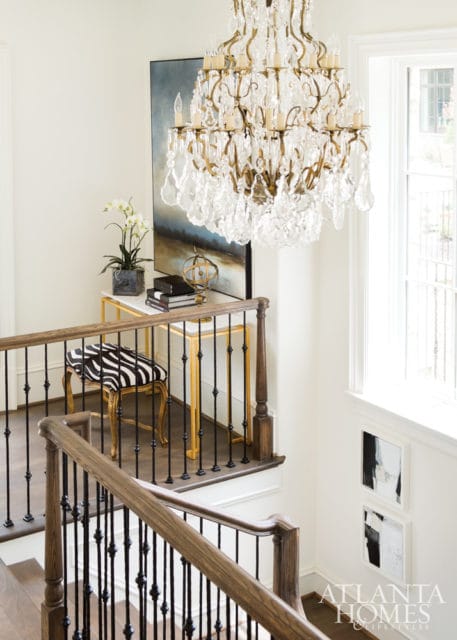 Staircase landing with a crystal chandelier, a gold console table with books and a zebra-print stool, and abstract art on the wall.