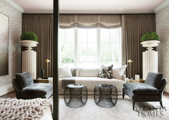 Living room with a white sofa, two dark upholstered chairs, black wire coffee tables, and large fluted pedestals with greenery. The room features patterned wallpaper and floor-to-ceiling brown curtains.