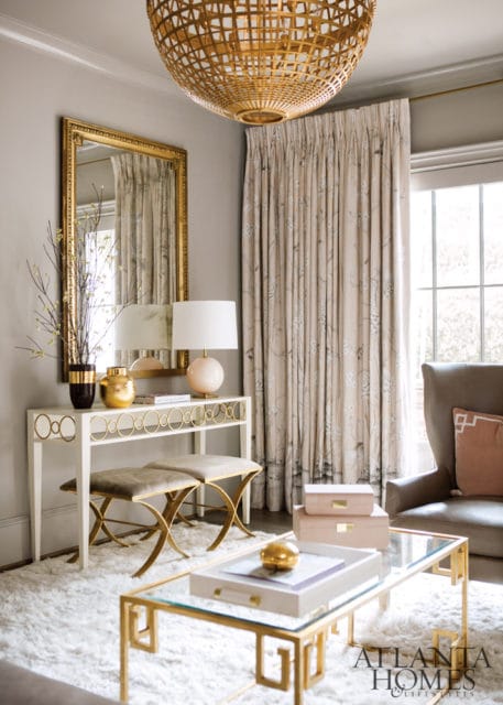 Living room with a gold pendant light, floral curtains, a white console table with gold accents, two upholstered stools, and a glass coffee table with gold frame.