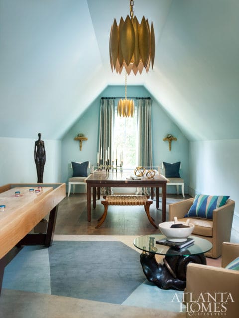 Light blue room with vaulted ceiling, featuring a shuffleboard table, a dark wood dining table with benches, two armchairs, and a gold lotus-shaped chandelier.