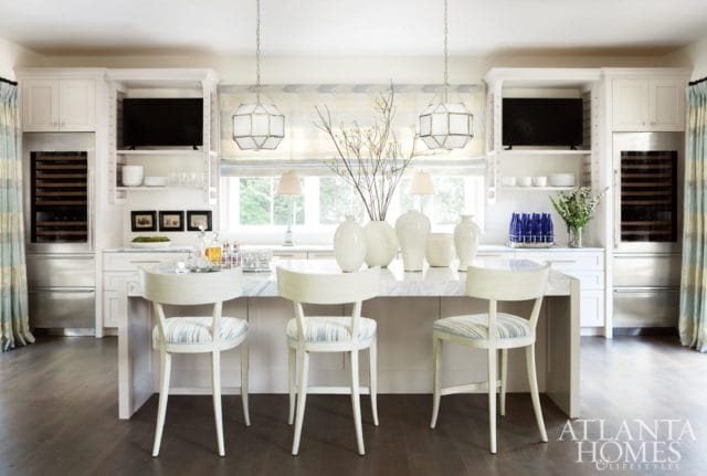 White kitchen island with marble countertop, three white chairs with striped cushions, and a collection of white vases. Built-in cabinetry with wine coolers and televisions flanks the space.