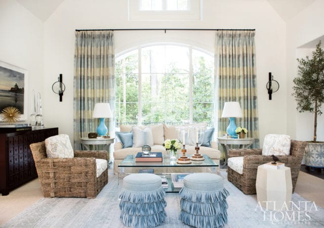 Living room with a cream sofa, woven armchairs, blue fringed ottomans, and a glass coffee table. Striped curtains frame a large arched window overlooking trees.
