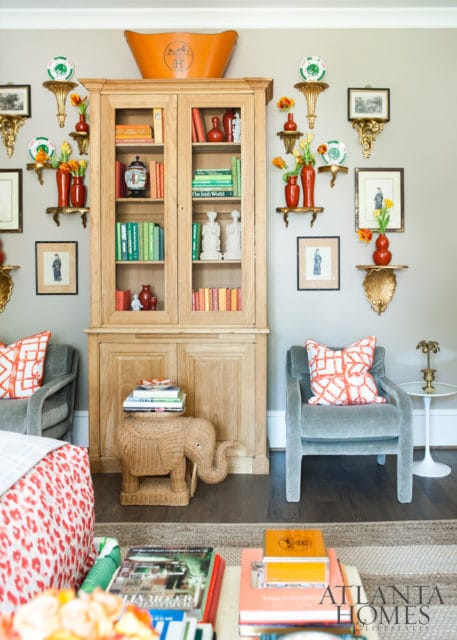 Living room with a light wood display cabinet filled with books and decorative items, flanked by gray armchairs with orange patterned pillows.
