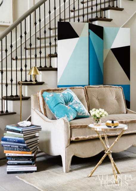 Beige tufted sofa with teal floral pillows next to a stack of books and a small round table with flowers and a decorative box. A geometric screen stands behind the sofa, with a staircase to the left.