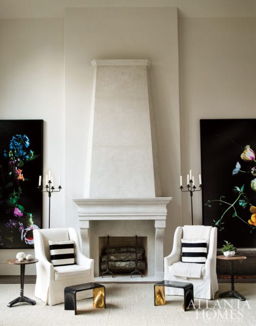 Living room with a tall, light stone fireplace, two white armchairs with striped pillows, and large floral artwork on black backgrounds.