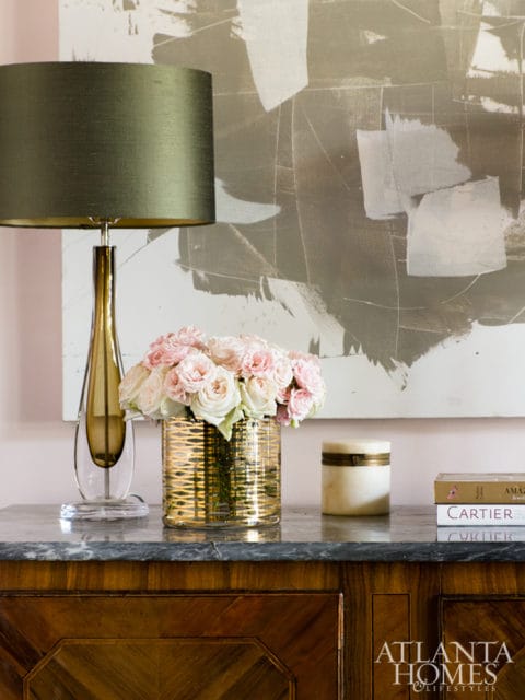 A table with a green lampshade, a vase of pink and white roses, a marble box, and books on a marble top dresser.