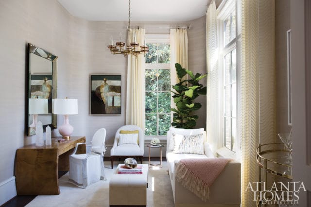 Light-filled living room with textured wallpaper, a wooden desk with a pink lamp and modern mirror, white chairs, a chaise lounge with a pink throw, and a large fiddle-leaf fig plant.
