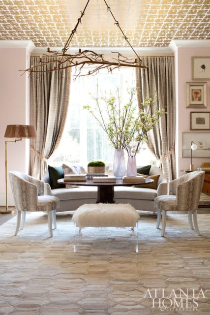 Living room with pale pink walls, gold patterned ceiling, and a branch chandelier. Features a curved sofa, two patterned armchairs, a round coffee table with vases and books, and a white fur ottoman.