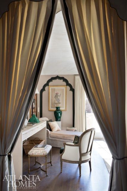 Interior view of a room with a white desk, a chaise lounge, and a white armchair, framed by draped curtains. A green vase and artwork are visible on the wall.