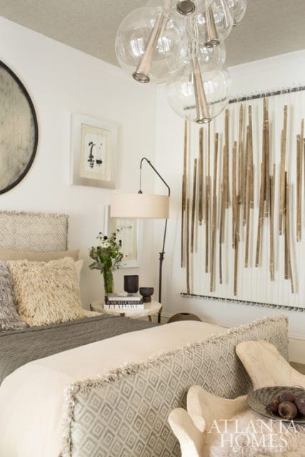 Bedroom with a grey upholstered headboard, textured pillows, and a large wall hanging made of vertical wooden sticks. A modern chandelier with glass globes hangs from the textured ceiling.