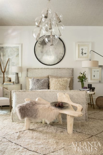 Bedroom with a patterned upholstered bed, textured pillows, a glass bubble chandelier, and a rustic wooden bench topped with sheepskin and fruit.