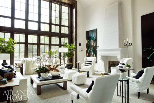 Living room with high ceilings, large grid windows, white sofas, white armchairs with black striped pillows, white fluffy ottomans, and a white fireplace.