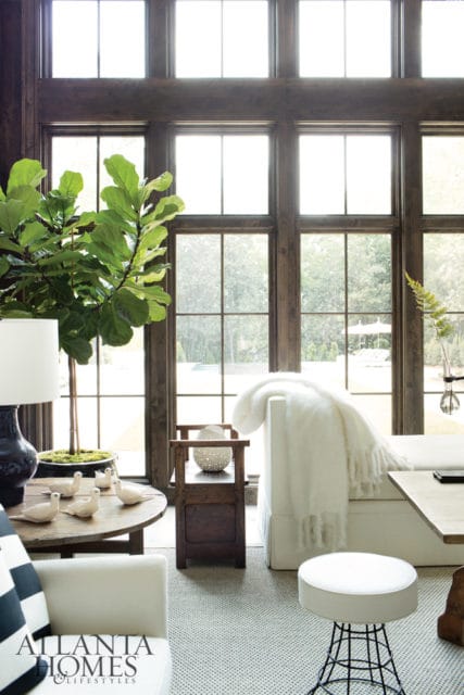 Living room with large windows, dark wood trim, a fiddle leaf fig plant, white sofa with a fluffy throw, and a round wooden coffee table with decorative birds.