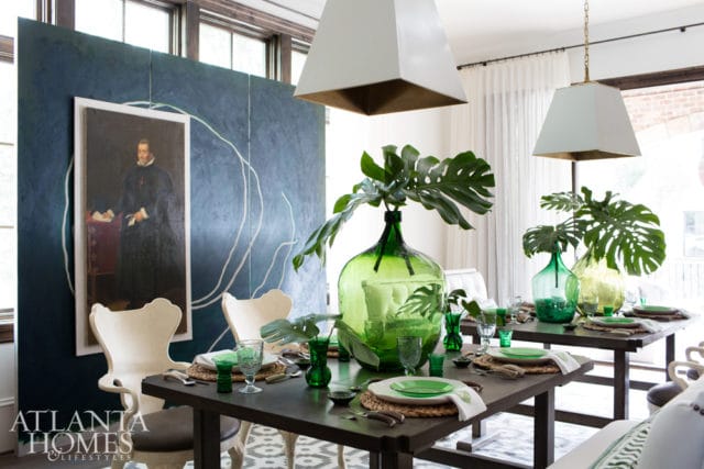 Dining room with two dark wood tables set for meals, featuring green glassware, woven placemats, and large green glass vases with monstera leaves. A large dark blue wall panel with a white abstract design hangs behind two cream upholstered chairs.