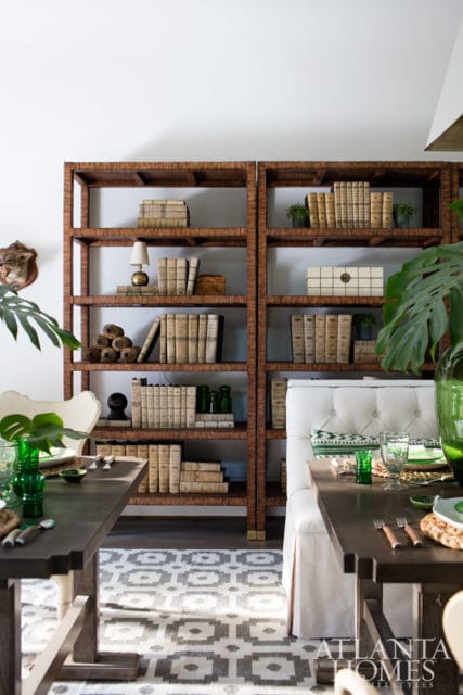 Dining room with dark wood tables, a large rattan bookshelf filled with books, and a tufted white bench. Green glassware and monstera leaves add pops of color.